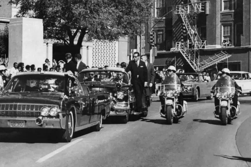 Vista a través del parabrisas del descapotable en primer plano, la mano del presidente John F. Kennedy se dirige hacia su cabeza pocos segundos después de recibir un disparo mortal, mientras la primera dama Jacqueline Kennedy le sujeta el antebrazo mientras la caravana avanza por Elm Street pasando por el Texas School Book Depository en Dallas, el 22 de noviembre de 1963. (James W. «Ike» Altgens/Foto AP)