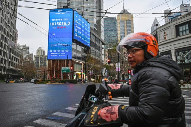 Un hombre pasa en scooter junto a una pantalla en la que se muestran las cifras del producto interior bruto en una calle de Shanghái el 19 de enero de 2026. Según datos oficiales, la economía china creció el año pasado a uno de los ritmos más lentos de las últimas décadas, en un contexto de gasto de consumo persistentemente bajo y de crisis de deuda en el sector inmobiliario del país. (Jade Gao/AFP vía Getty Images).